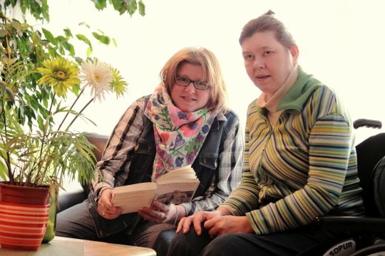Eine Alltagshelferin liest gemeinsam mit einer im Rollstuhl sitzenden jungen Frau aus einem Buch.