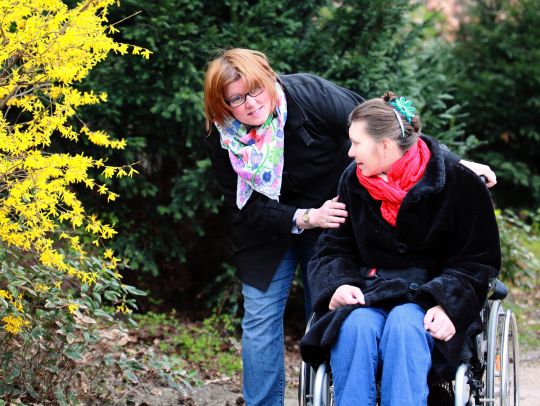 Eine Alltagshelferin schiebt eine im Rollstuhl sitzende junge Frau durch eine Parkanlage.