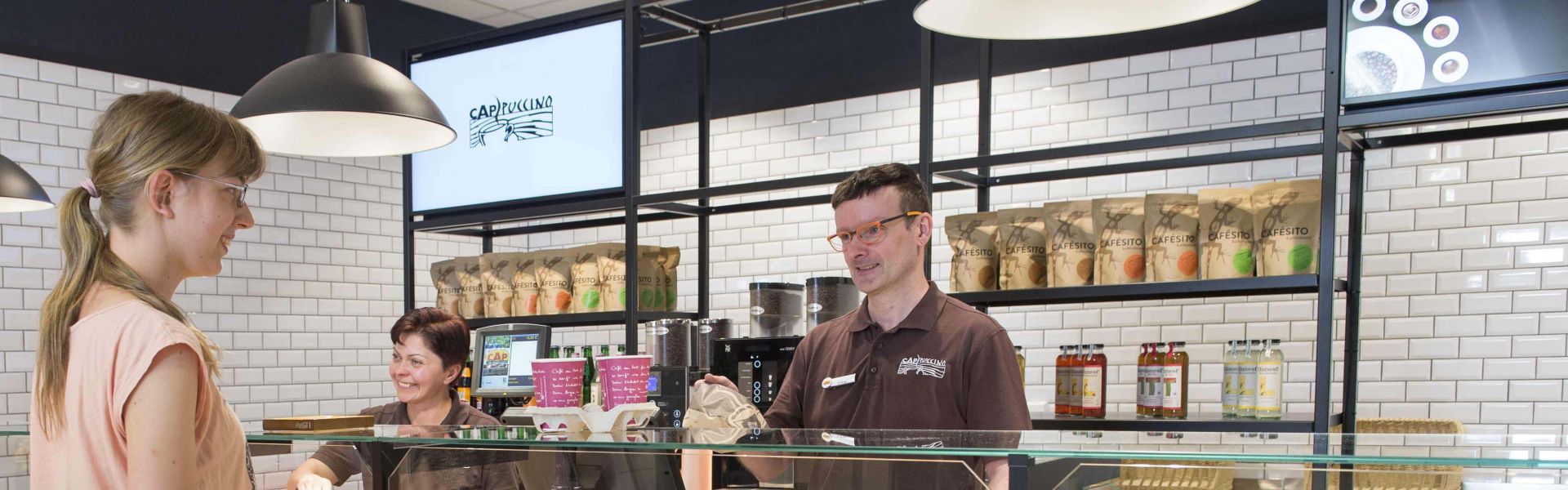 Zwei Mitarbeitende des Backshops Cappuccino hinter der Theke, sie bedienen eine Kundin.