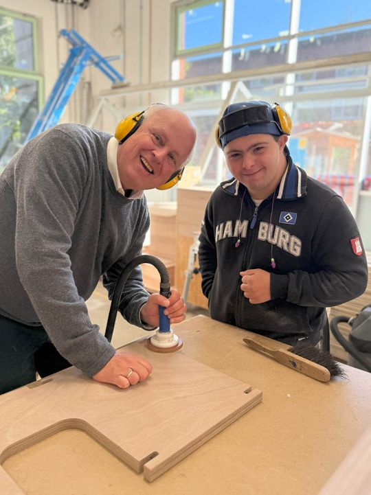 Ein Herr mittleren Alters und ein jüngerer Mann lächeln in die Kamera. Sie machen Holzschleifarbeiten in einer Werkstatt