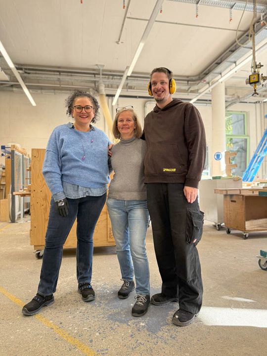 Zwei Frauen mittleren alters, und ein hoch gewachsener Mann posieren stehend in der Holzwerkstatt