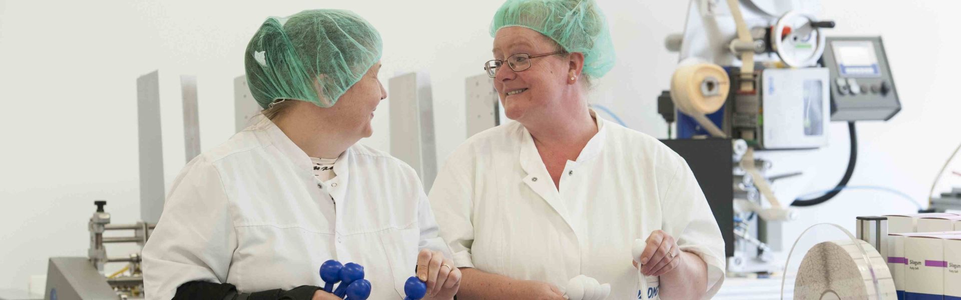 Zwei Frauen sitzen an einem Tisch. Sie schauen sich an. Beide tragen weiße Kittel und haben eine grüne Haube auf dem Kopf.