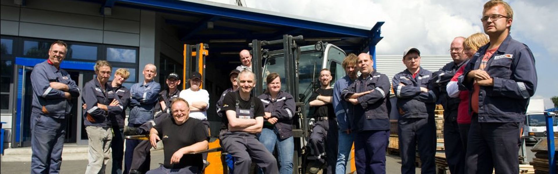 Größere Gruppe Mitarbeitender in Arbeitskleidung posieren vor einem Gabelstabler.
