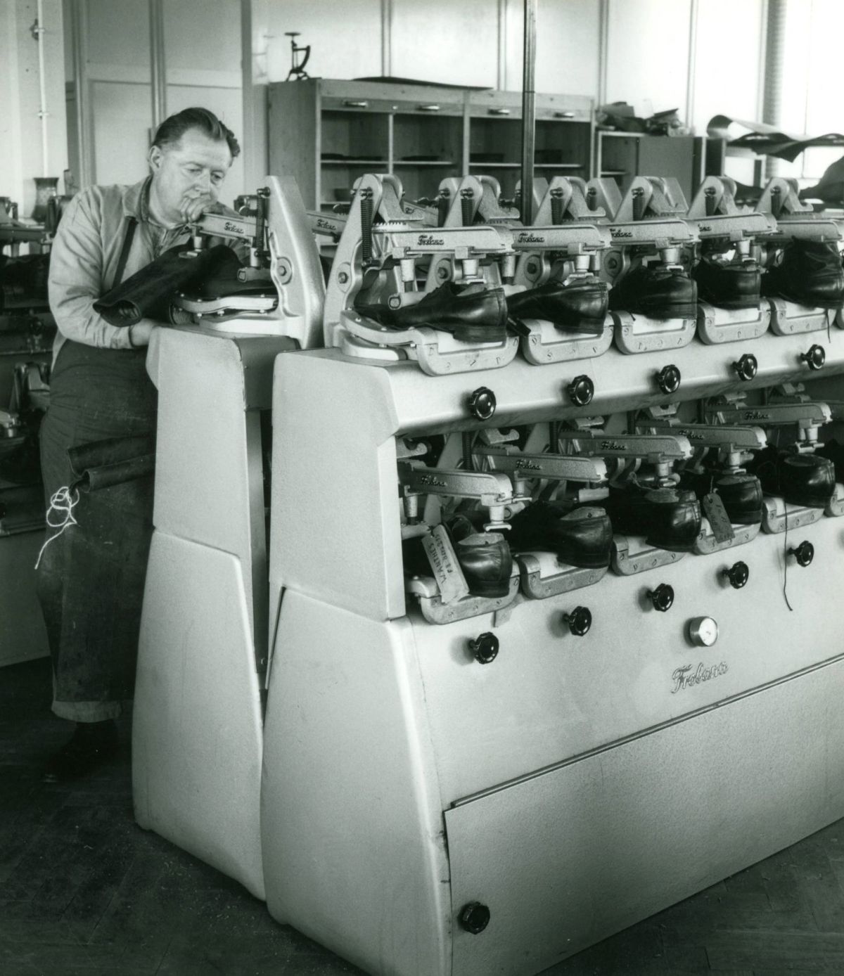 Es ist ein altes schwarz-weiß-Foto. Auf dem Foto steht ein Mann an einer Maschine und repariert Schuhe.