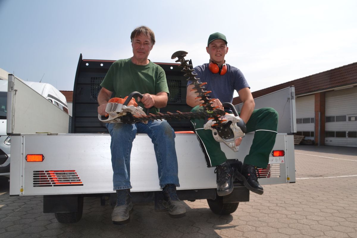 Zwei Mitarbeiter der Arbeitsgruppe Gartenbau sitzen am Ende der Ladefläche eines LKWs. Sie halten elektrische Heckenscheren in den Händen.