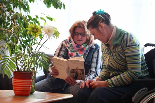 Eine Alltagshelferin liest gemeinsam mit einer jungen Frau in einem Buch.