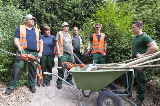Die Arbeitsgruppe Gartenbau neben einem großen Schubkarren voller Arbeitsgeräten. Sie tragen grüne Arbeitshosen und orangefarbene Westen.