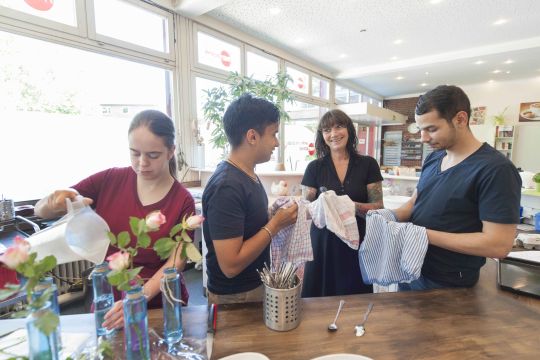 Eine Gruppe Gastronomiemitarbeitender beim Arbeiten in einer Küche. Eine Kollegin befüllt Blumenvasen, drei weitere trocknen Geschirr.