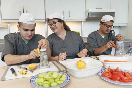 Mitarbeitende des Lehrcafés schneiden Gemüse an einer Küchenarbeitsfläche. Sie tragen graue Kochhemden und weiße Mützen.