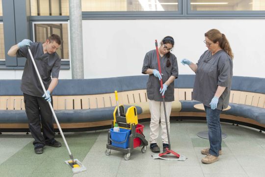 Mitarbeitende des Hauswirtschaft und Lehrcafés wischen mit Wischmopps den Boden in einem Wartebereich.