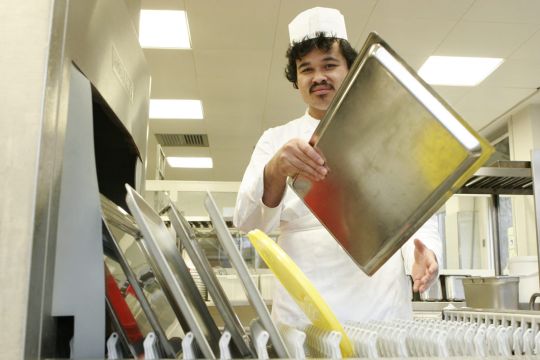Ein Mitarbeiter in Küchenarbeitskleidung stapelt Tabletts in eine industrielle Spülmaschine.