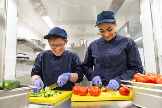 Zwei Frauen stehen an einem Küchentresen. Sie schneiden Gemüse mit Messern. 