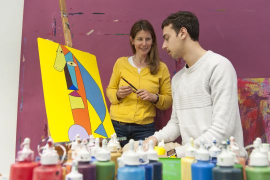 Zwei Personen neben einem Gemälde auf einer Staffelei. Es ist in kräftigen Farben gemalt, der Hintergrund ist gelb.