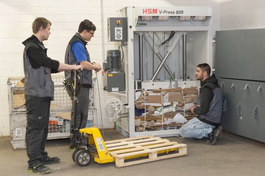 Mitarbeitende beim Entleeren einer Papp-Presse unterstützen sich gegenseitig mit einer Eruopalette und einem Hubwagen.